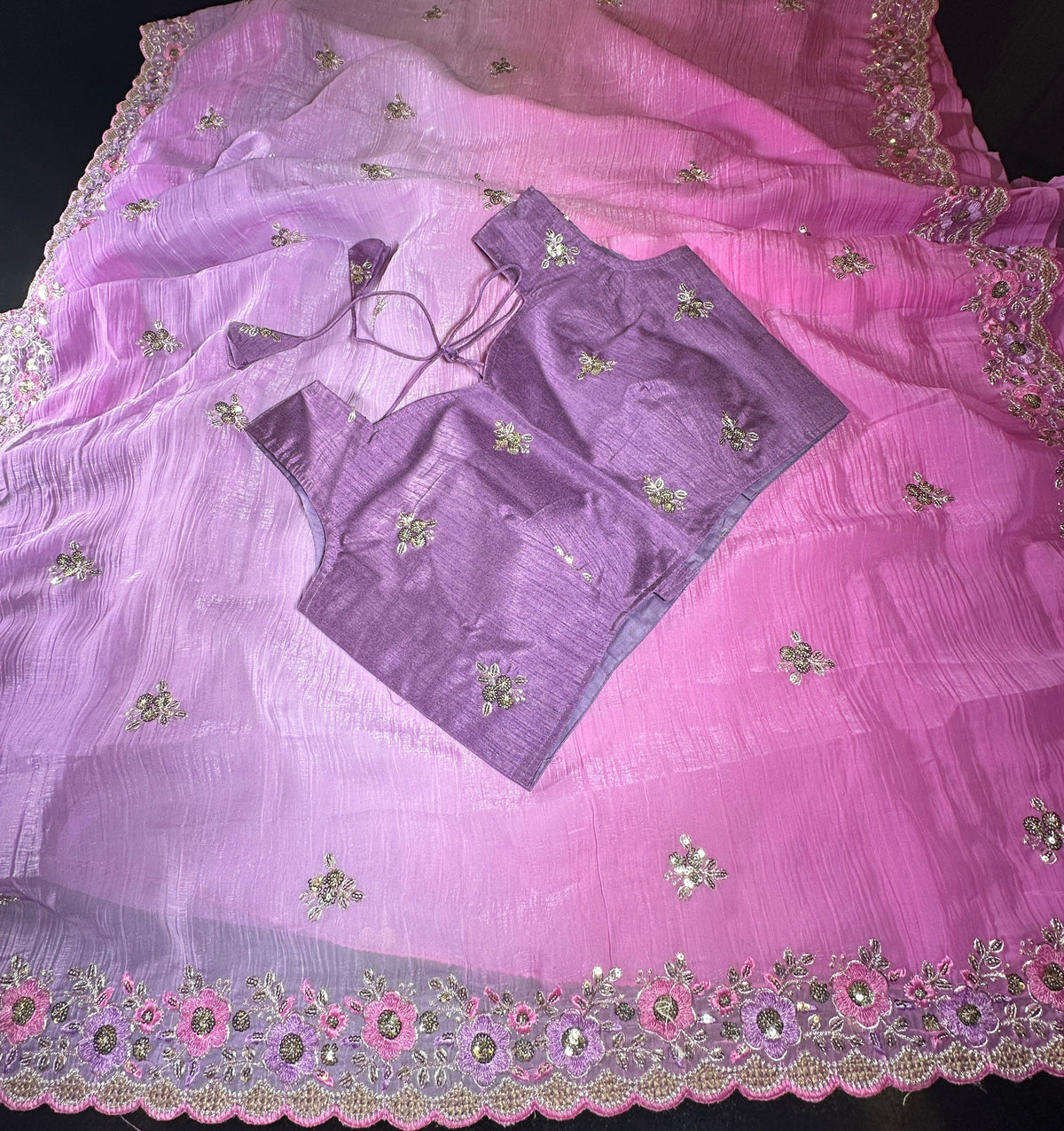 A pink and purple gradient saree with floral embroidery and mirror work along the border, laid flat on a dark surface with a matching purple blouse piece placed on top.