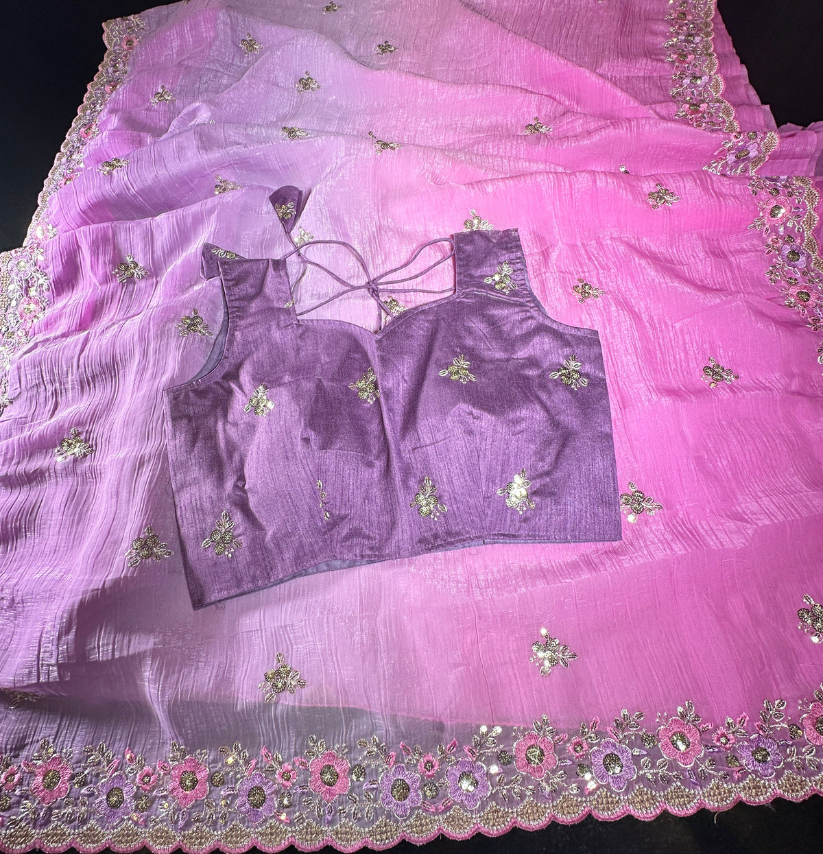A pink and purple traditional Indian outfit set, featuring a sheer pink dupatta with floral embroidery and mirror work, a matching purple blouse with tie-back straps, and a purple skirt, laid out on a dark surface.